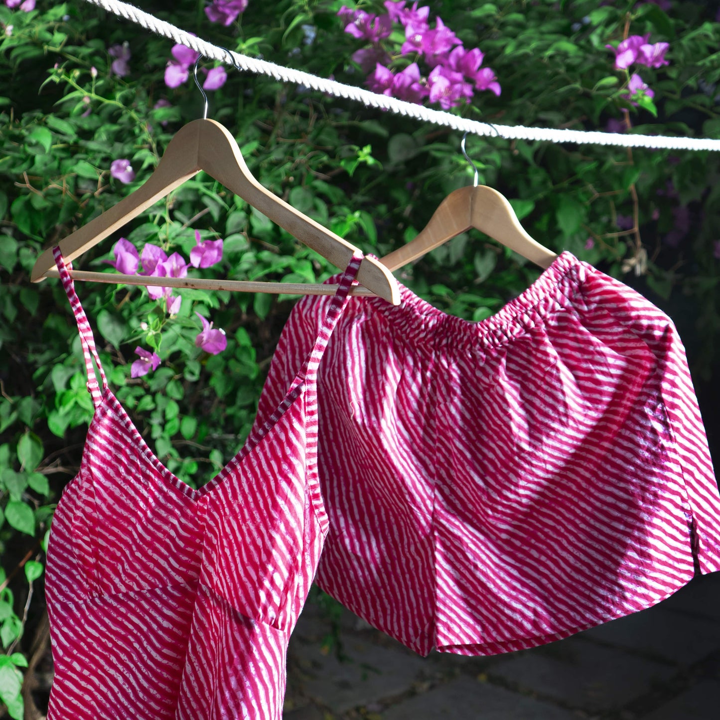 Pink striped cami set and shorts against a green bush background on a wooden hanger hanging on a white rope.